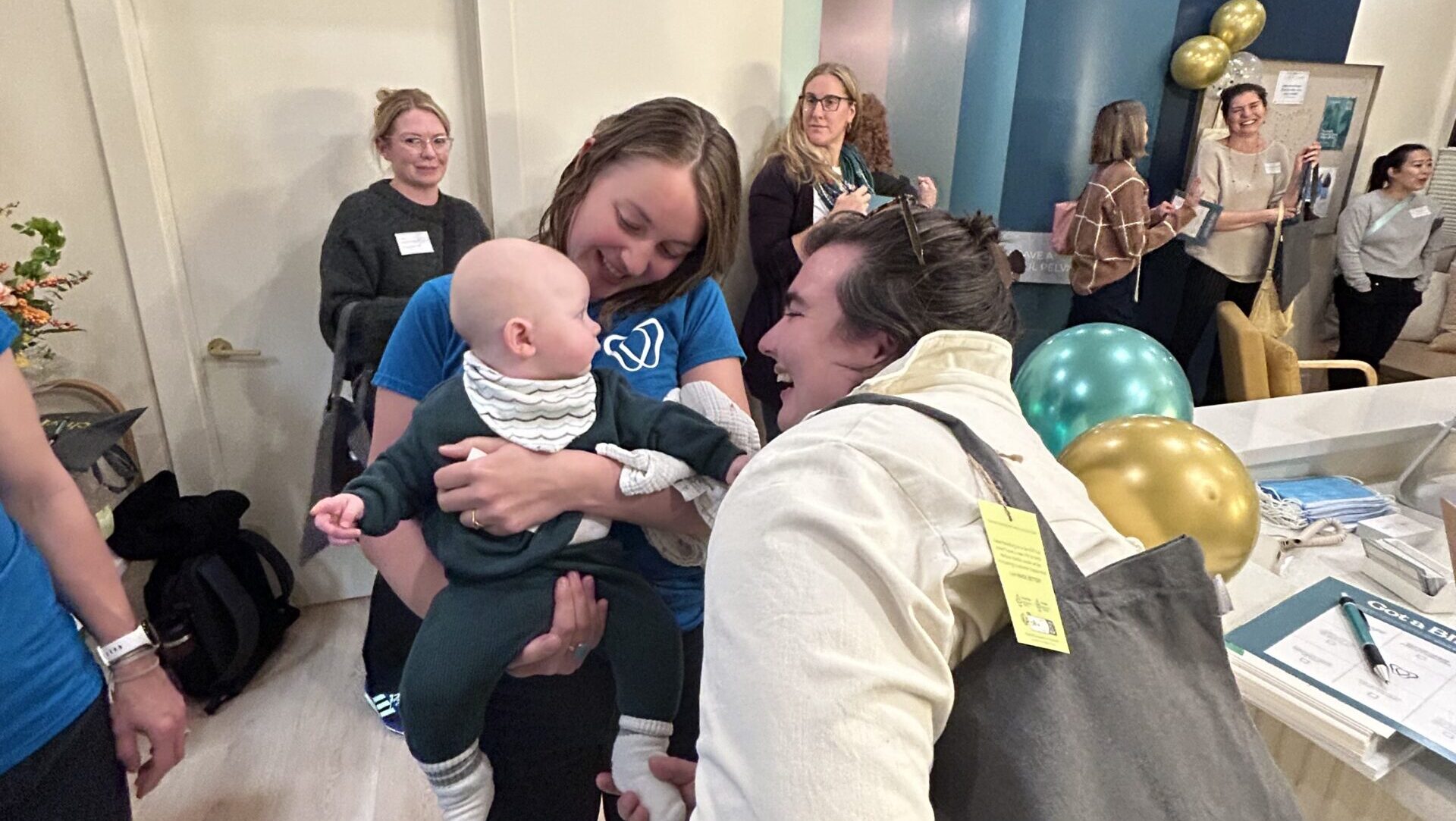 A candid shot from the launch party. Anniken holds a baby who is smiling at a guest holding a tote bag full of sponsored goodies. In the background, the party attendees smile and converse.