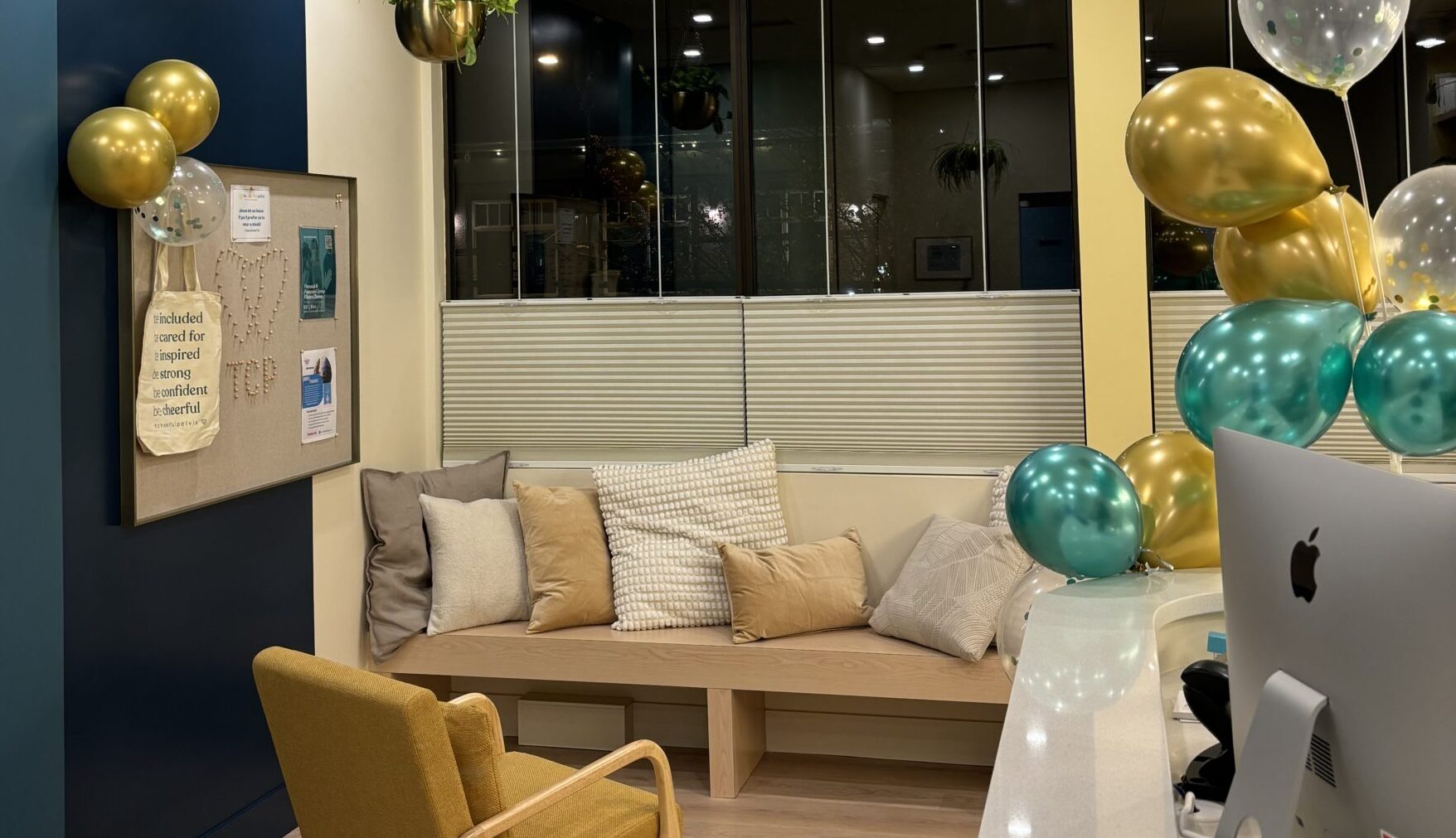 The new Cheerful Pelvis waiting room, filled with gold and turquoise balloons. There is a long bench covered with pillows by the window wall, and a reception desk with computer monitor to the right. 