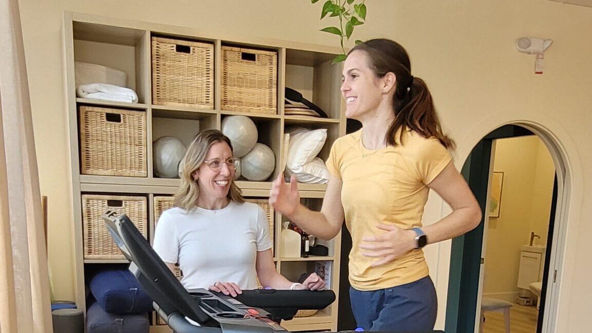 A woman coaching another woman as she runs on a treadmill, offering support and guidance during a workout session