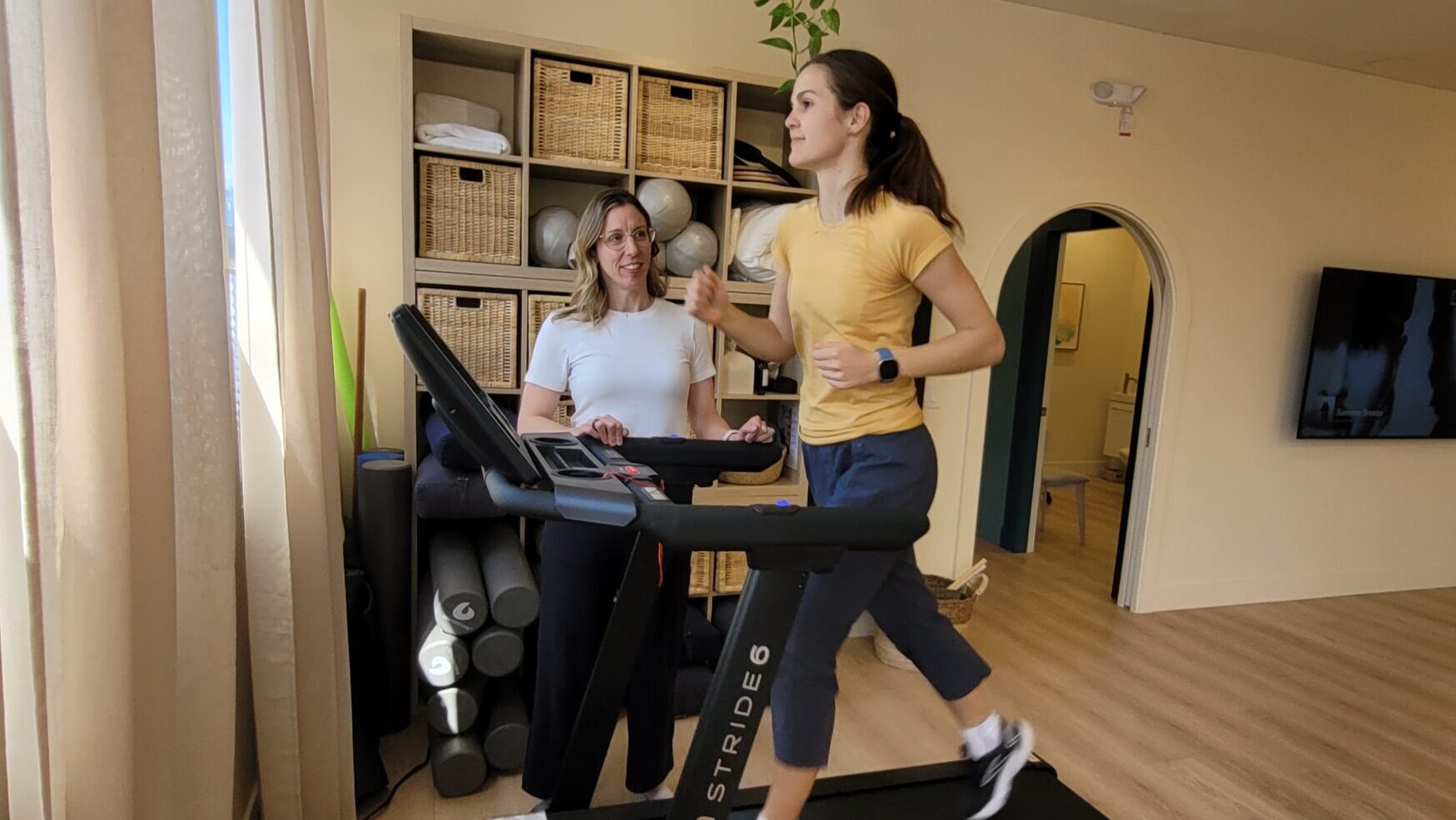 A woman coaching another woman as she runs on a treadmill, offering support and guidance during a workout session